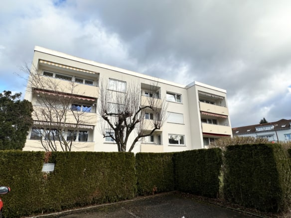 Ein dreistöckiges weißes Wohnhaus steht hinter hohen grünen Hecken und blattlosen Bäumen unter einem bewölkten Himmel. Perfekt für diejenigen, die eine Wohnung verkaufen Ravensburg, mehrere Balkone und Fenster sind auf der Fassade sichtbar.