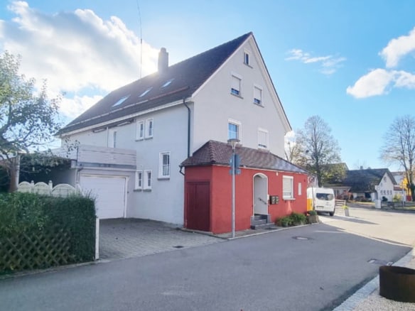 Ein hellgraues dreistöckiges Haus mit einem kleinen roten Anbau steht in einer ruhigen Straße in Ravensburg, perfekt für diejenigen, die mit einem erfahrenen Immobilienmakler Ravensburg verkaufen wollen. Bäume und Nachbarhäuser umgeben es, ein weißer Lieferwagen ist in der Nähe geparkt.