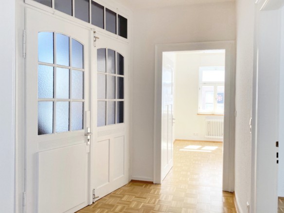 Helle Diele mit weißen Türen, Parkettboden und Milchglasscheiben. Sonnenlicht strömt aus einem angrenzenden Raum herein und schafft eine klare, luftige Atmosphäre - ideal für alle, die eine Wohnung mit Stil und Komfort in Ravensburg verkaufen möchten.
