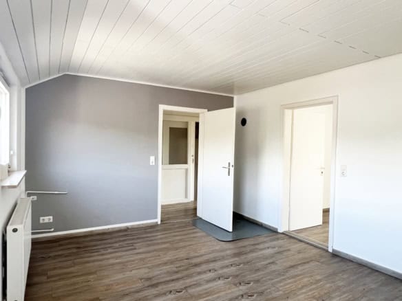 Ein sauberer, leerer Raum mit hellgrauen Wänden, Holzfußboden und weißer Decke - ideal, wenn Sie eine Wohnung in Ravensburg verkaufen möchten. Zwei offene Türen und ein Fenster mit natürlichem Licht werten den Raum auf; ein Heizkörper und eine graue Matte setzen einfache Akzente.