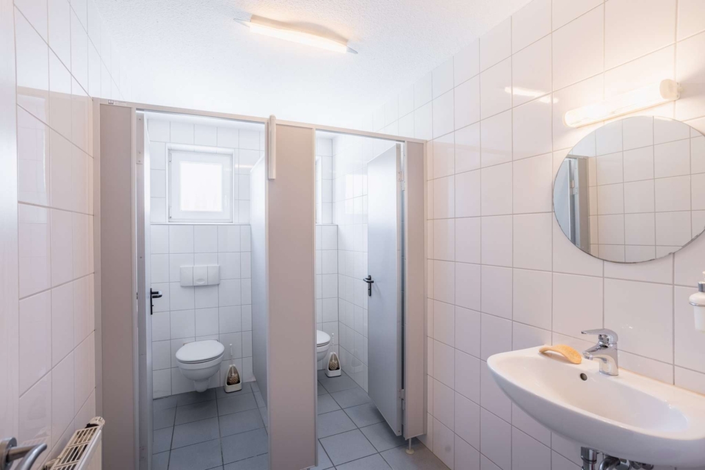 Eine saubere öffentliche Toilette mit zwei Toilettenkabinen, jeweils mit einer weißen Toilette und einer Tür, weiß gefliesten Wänden, einem runden Spiegel über einem weißen Waschbecken und einem Fenster, das für Tageslicht sorgt.