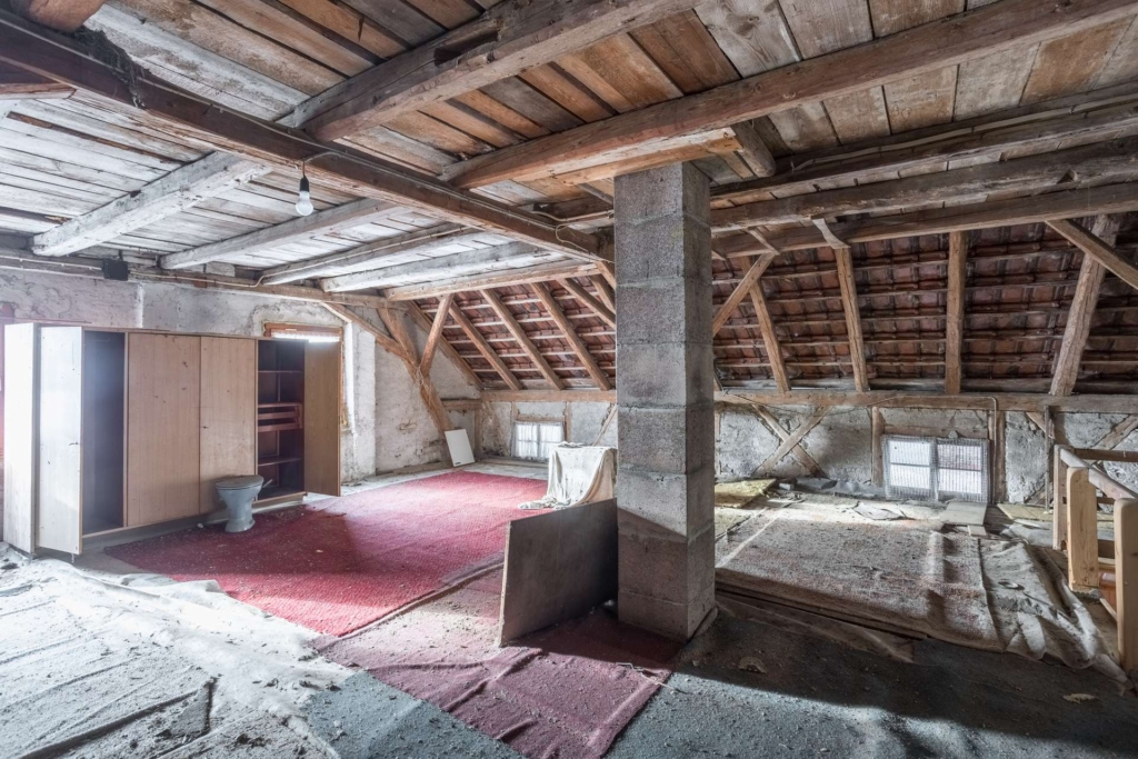 Ein alter Dachboden mit freiliegenden Holzbalken, einer Betonsäule, rotem Teppich, einem Kleiderschrank, einer Toilette und verstreuten Trümmern. Das Sonnenlicht fällt durch kleine Fenster und beleuchtet das abgenutzte und staubige Innere.