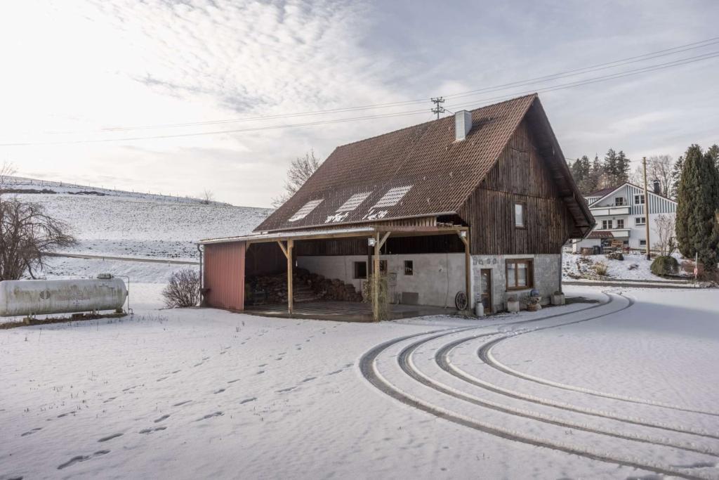 Eine rustikale Holzscheune mit einem roten Ziegeldach steht in einer verschneiten Landschaft. Reifenspuren schlängeln sich durch den Schnee, und Fußspuren führen zur Scheune. Im Hintergrund sind ein Hügel und Häuser unter einem bewölkten Himmel zu sehen.