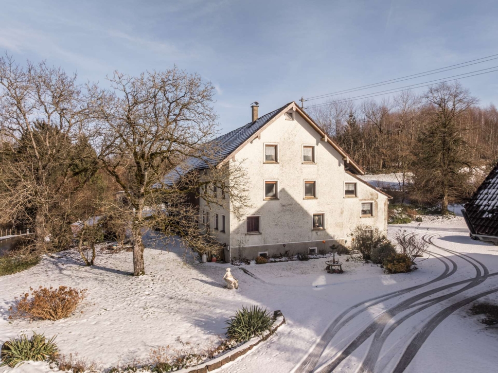 Ein weißes, zweistöckiges Haus mit schrägem Dach steht inmitten einer verschneiten Landschaft, umgeben von kahlen Bäumen und Sträuchern. Reifenspuren schlängeln sich über die schneebedeckte Einfahrt, und das Gelände ist von Wald umgeben.