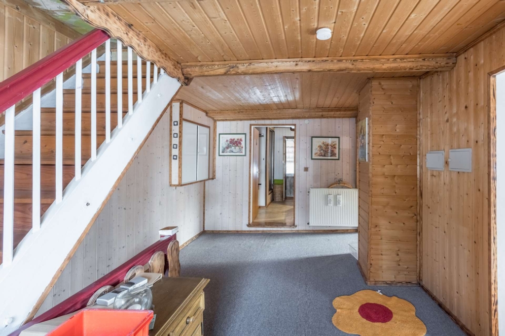 Ein gemütlicher Flur mit holzgetäfelten Wänden und Decken, Teppichboden, einer Treppe mit rotem Geländer, Wandkunst, einem Heizkörper und einem blumenförmigen Teppich neben einer Holztür.