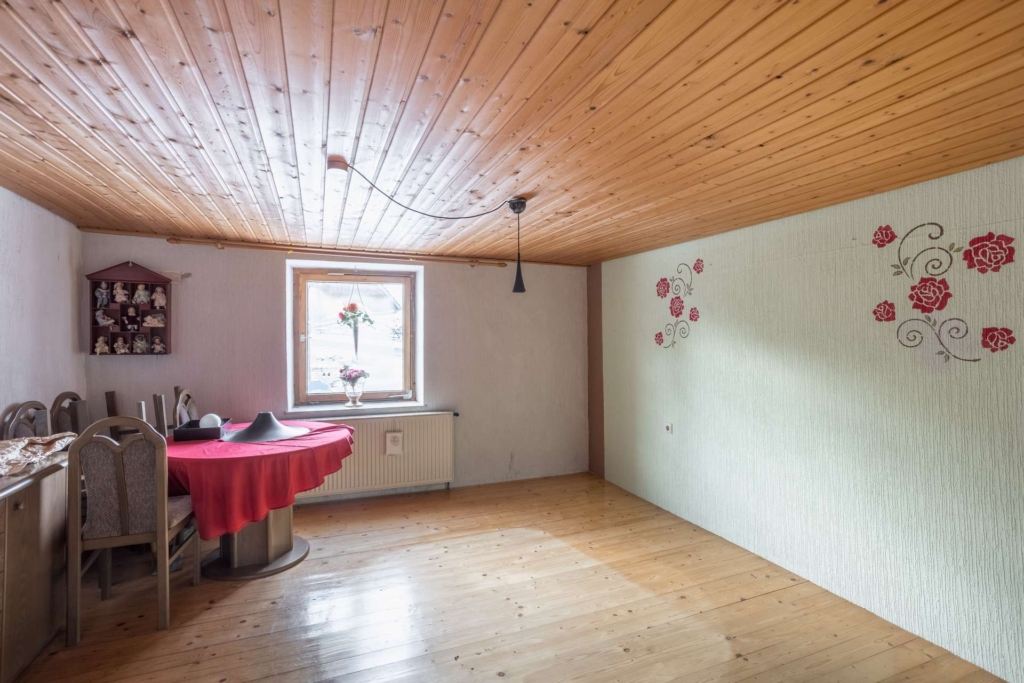 Ein kleiner, heller Raum mit Holzboden und -decke, einem mit einem roten Tuch bedeckten Tisch, mehreren Stühlen, einem Fenster, das Tageslicht hereinlässt, einem Wandregal mit Figuren und floralen Wandbildern.