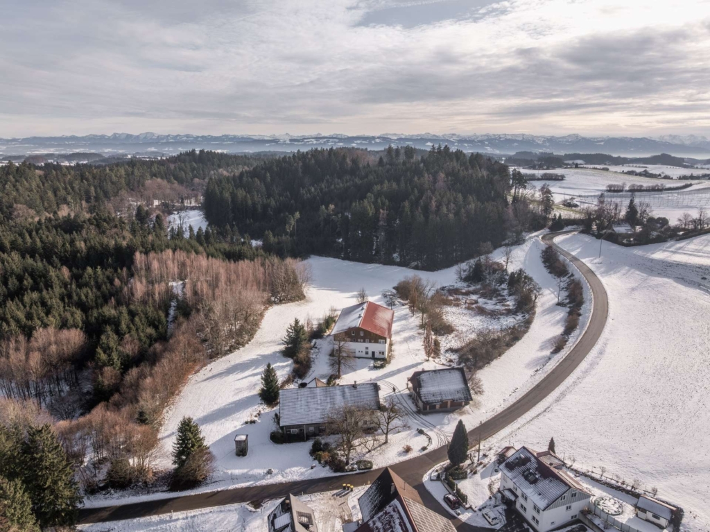 Luftaufnahme einer ländlichen Landschaft im Winter mit schneebedeckten Feldern, verstreuten Häusern, einer kurvenreichen Straße und einem dichten Wald unter einem bewölkten Himmel.