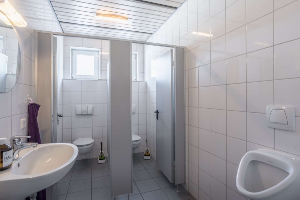 Eine saubere öffentliche Toilette mit zwei Toilettenkabinen, einem weißen Waschbecken mit Seifenspender, einem Urinal, gefliesten Wänden und Böden sowie zwei kleinen Fenstern, die Tageslicht hereinlassen.