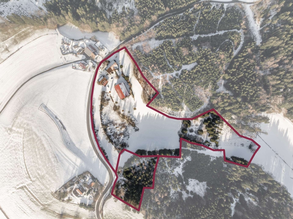 Luftaufnahme einer verschneiten ländlichen Landschaft mit Gebäuden und Waldgebieten. Ein roter Umriss markiert einen bestimmten, unregelmäßig geformten Landabschnitt, der einen Teil des Waldes und mehrere Gebäude umfasst.