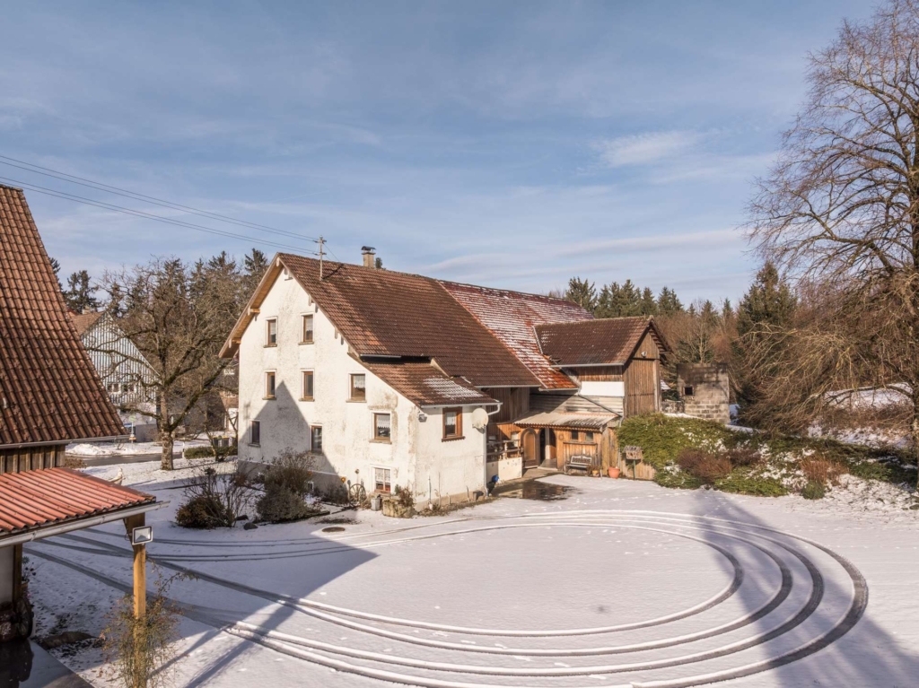 Ein weißes Bauernhaus mit rotem Dach steht in einem verschneiten Hof, umgeben von Bäumen. Im Schnee sind geschwungene Reifenspuren zu erkennen, und über dem Haus hängt ein blauer Himmel.