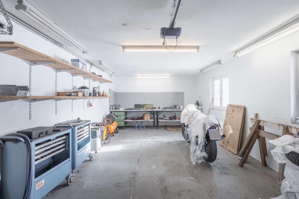 Eine saubere, geräumige Garage mit weißen Wänden, Werkbänken im hinteren Bereich, Regalen mit Werkzeugen, zwei blauen Werkzeugschränken auf der linken Seite und einem mit Plastik verkleideten Motorrad in der Mitte. Leuchtstoffröhren hängen von der Decke.
