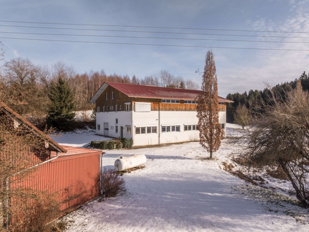 Ein großes Gebäude mit rotem Dach und weißen unteren Wänden steht in einer verschneiten Landschaft, umgeben von kahlen Bäumen und einigen Immergrünen. Im Vordergrund sind ein Propantank und ein weiteres Gebäude mit rotem Dach zu sehen.