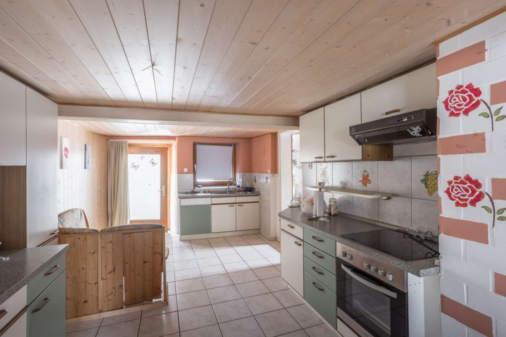 Eine gemütliche Küche mit weißen Schränken, gefliestem Boden, Elektroherd, Backofen und Dunstabzugshaube. Die Holzdecke und die Vertäfelung sorgen für eine warme Atmosphäre. Dekorative rote Rosenfliesen akzentuieren die Wand. Durch ein Fenster und eine Glastür fällt natürliches Licht ein.