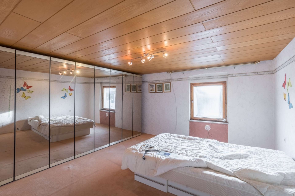 Ein Schlafzimmer mit heller Holzdecke, verspiegelten Schranktüren an einer Wand, einem Bett mit weißer Bettdecke, zwei Fenstern, Schmetterlings-Wandbildern und gerahmten Bildern über dem Bett, alles in sanften, neutralen Tönen.