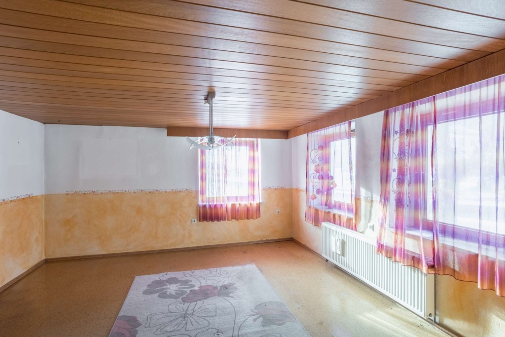 Ein leerer Raum mit holzgetäfelter Decke, weißen und orangefarbenen Wänden, zwei Fenstern mit durchsichtigen lila Vorhängen, einem Heizkörper unter den Fenstern, einer modernen Hängelampe und einem Blumenteppich auf dem Boden.