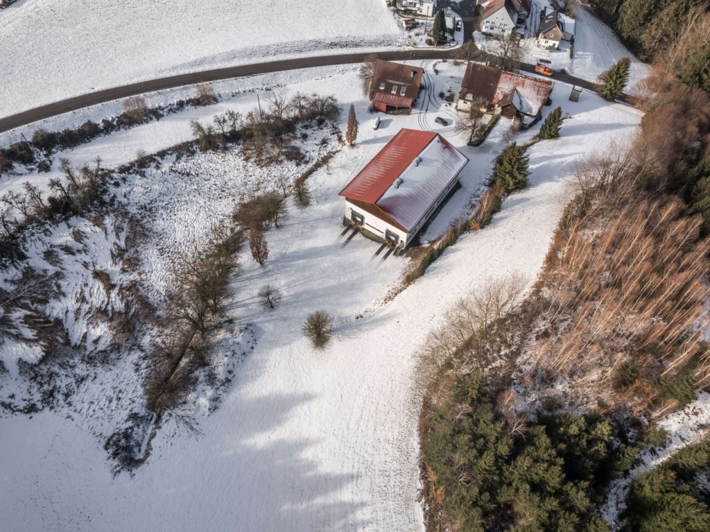 Luftaufnahme einer ländlichen, verschneiten Landschaft mit einem großen Gebäude mit rotem Dach, umgeben von Bäumen und Feldern. Einige Häuser und eine Straße sind in der Nähe des oberen Bildrandes zu sehen.