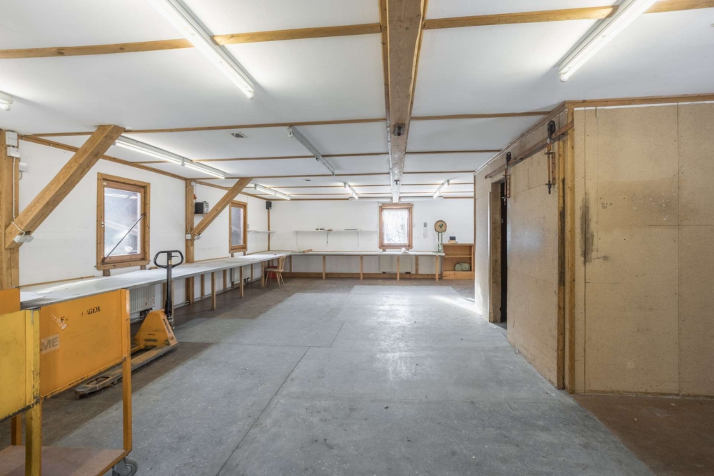 Eine geräumige, gut beleuchtete Werkstatt mit weißen Wänden, freiliegenden Holzbalken, Leuchtstoffröhren, langen Werkbänken, ein paar Fenstern, einer Uhr an der Wand und Geräten wie einem gelben Palettenheber.