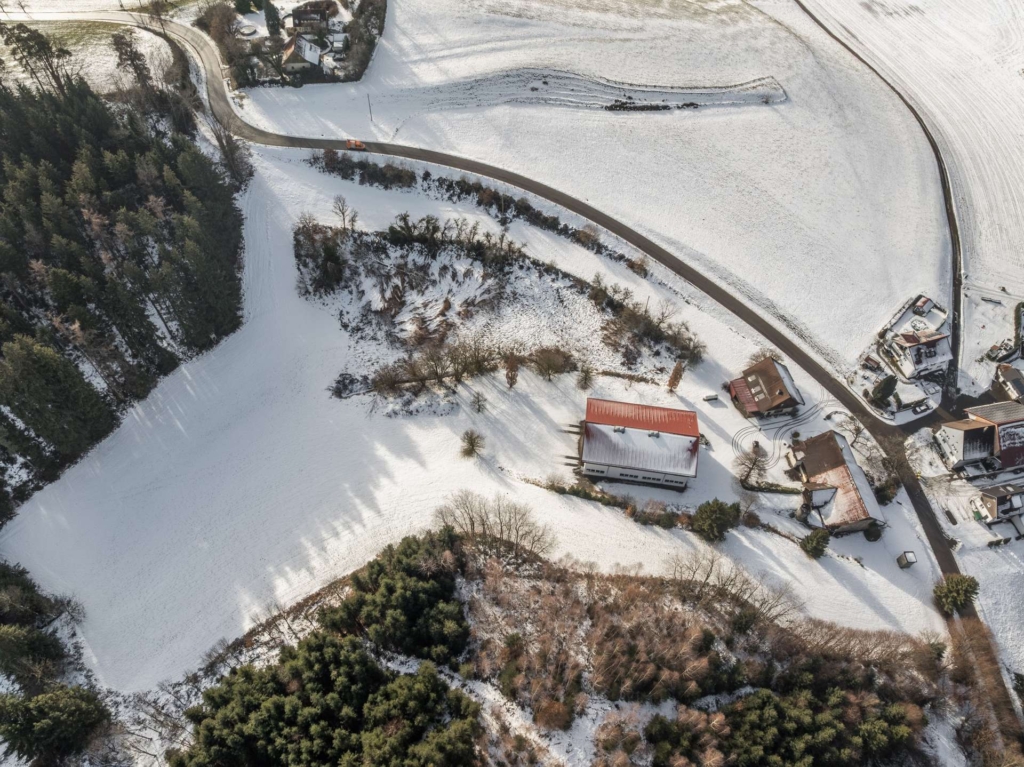 Luftaufnahme von schneebedeckten Feldern, verstreuten Häusern mit roten Dächern, kurvenreichen Straßen und Waldstücken in einer ländlichen Winterlandschaft.