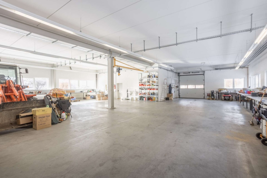 Geräumige, gut beleuchtete Industriewerkstatt mit Betonboden, Werkbänken an der rechten Wand, Regalen mit geordneten Vorräten, großen Fenstern und einem teilweise sichtbaren orangefarbenen Traktor auf der linken Seite.