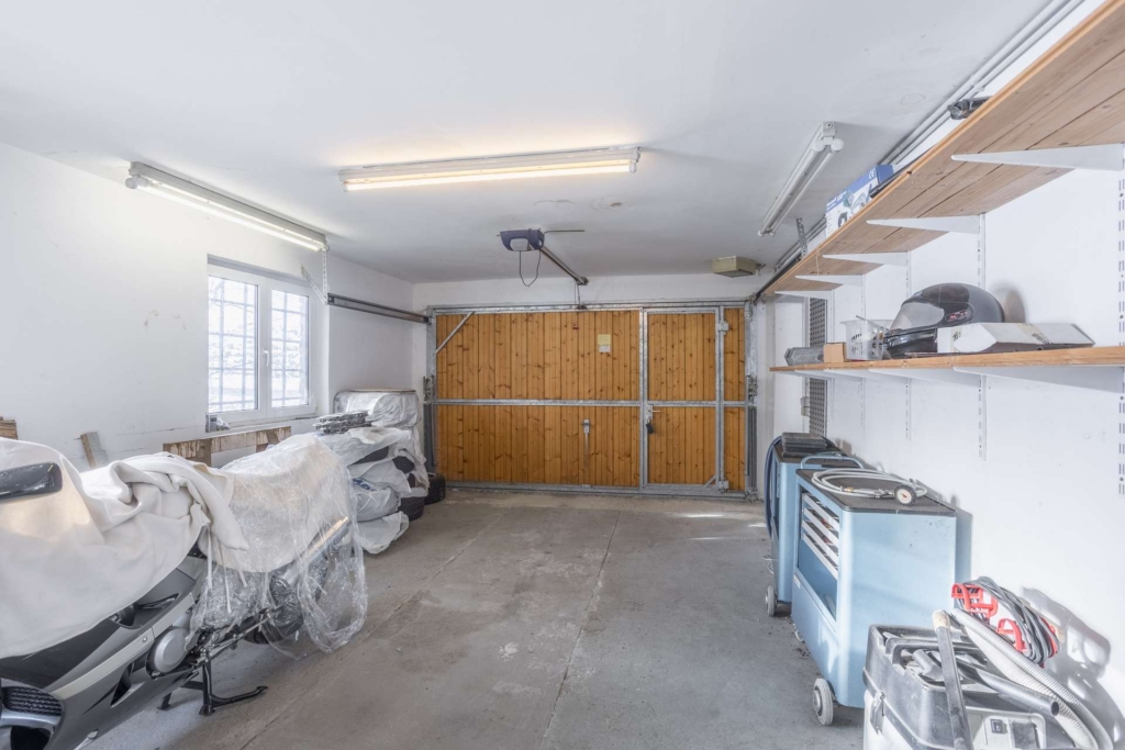 Eine saubere, geräumige Garage mit einem hölzernen Garagentor, weißen Wänden, Oberlichtern, Regalen mit Werkzeugen und Geräten auf der rechten Seite und abgedeckten Gegenständen an der linken Wand neben einem Fenster.
