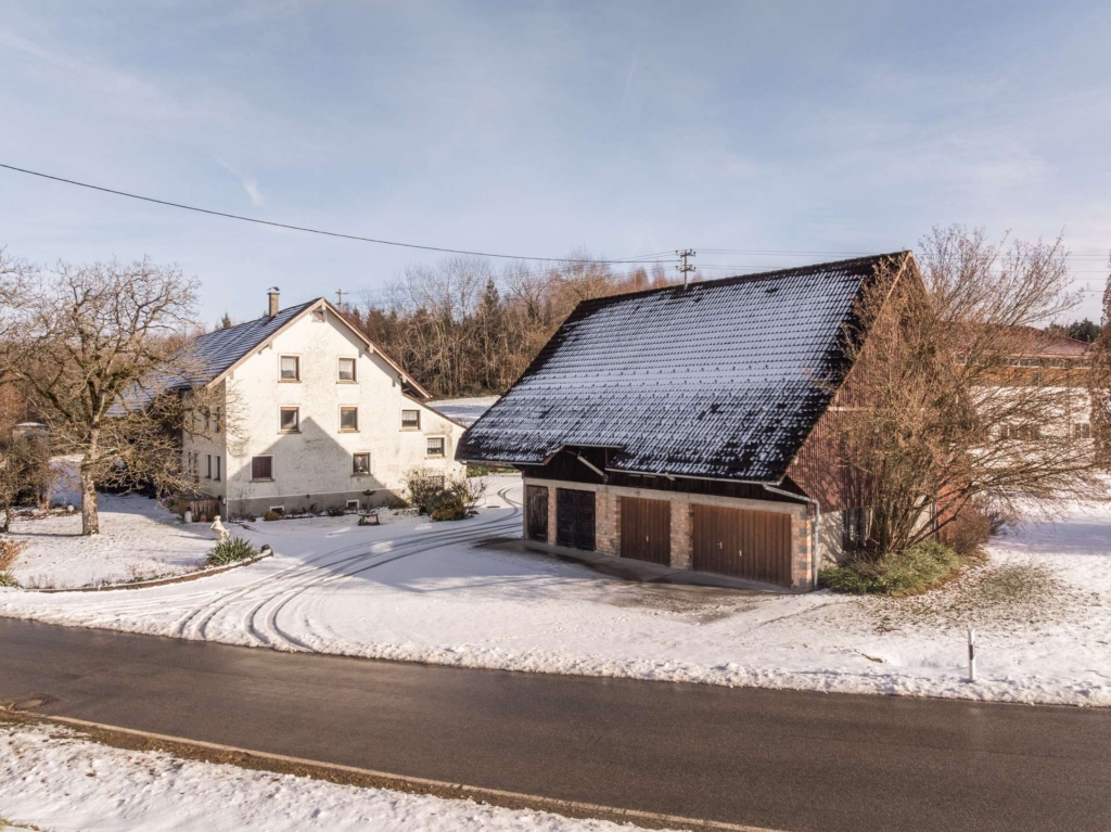 Eine schneebedeckte ländliche Szene mit einem weißen Haus und einer braunen Scheune, beide umgeben von Bäumen und Feldern. Im Schnee sind Reifenspuren zu erkennen, die von der Straße im Vordergrund zu den Gebäuden führen.