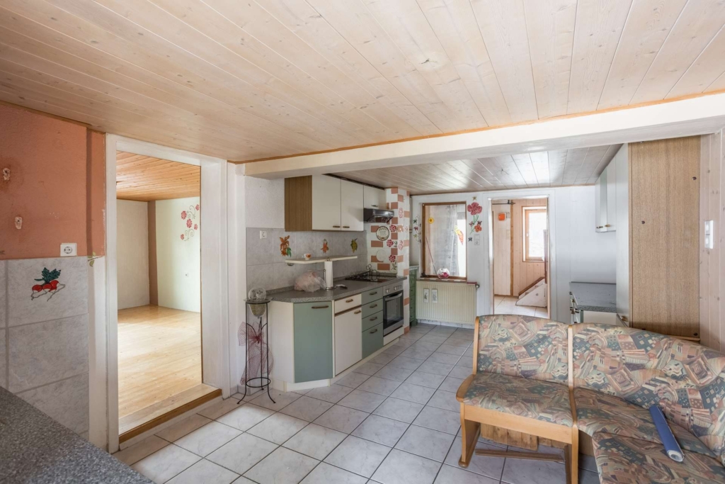 Eine kleine, veraltete Küche mit hellem Fliesenboden, holzgetäfelter Decke, pastellfarbenen Schränken und geblümten Wandbildern. Im Vordergrund steht eine gemusterte Couch; ein weiterer Raum mit Holzböden ist durch eine Türöffnung zu sehen.