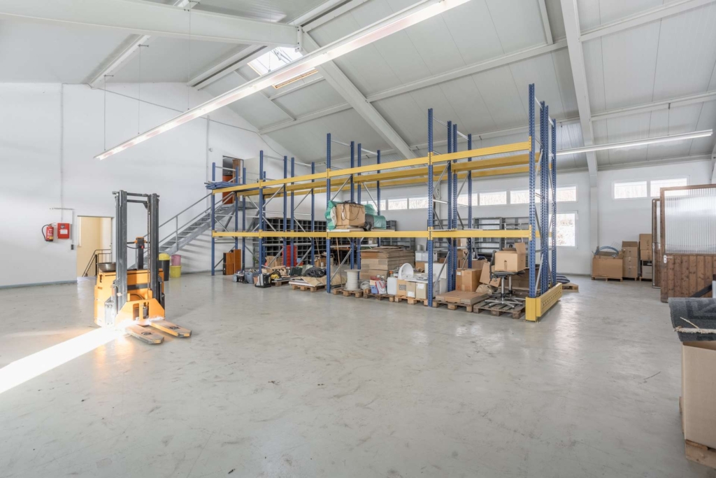 Geräumiges Lagerhaus mit hohen Decken, Lagerregalen mit verschiedenen Kisten und Geräten, einem gelben Gabelstapler auf der linken Seite und verstreuten Gegenständen auf dem Boden; durch die Fenster fällt Sonnenlicht.