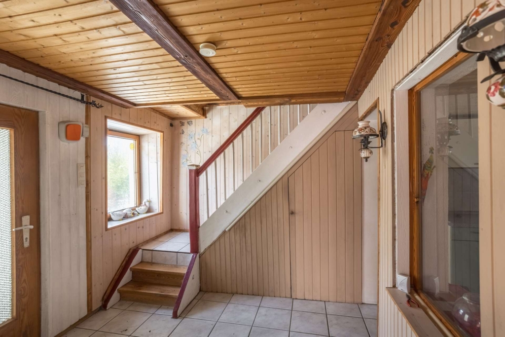 Ein kleiner Eingangsbereich mit gefliestem Boden, Holzwänden und einer Holzdecke. Eine Treppe mit weißer Verkleidung und braunen Zierleisten führt nach oben, und unter der Treppe befindet sich eine Tür. Fenster lassen natürliches Licht in den Raum.