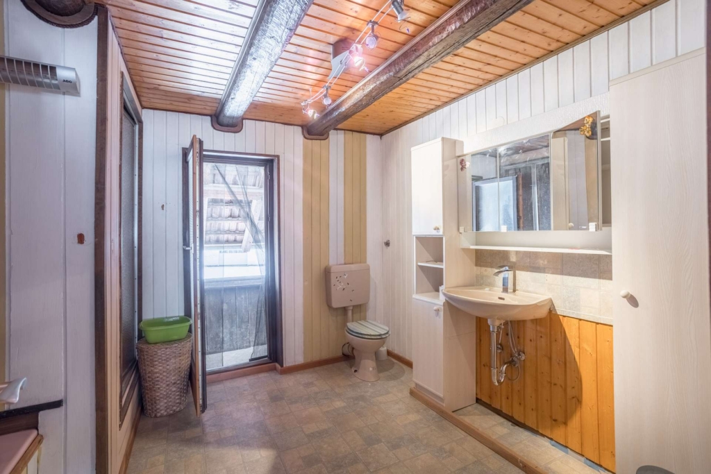 Ein rustikales Badezimmer mit holzgetäfelten Wänden und Decken, einer Toilette, einem Waschbecken mit Spiegel, offenen Regalen, einem Wäschekorb und einer Tür, die zu einem Außenbereich führt. An der Decke sind freiliegende Rohre und Balken zu sehen.