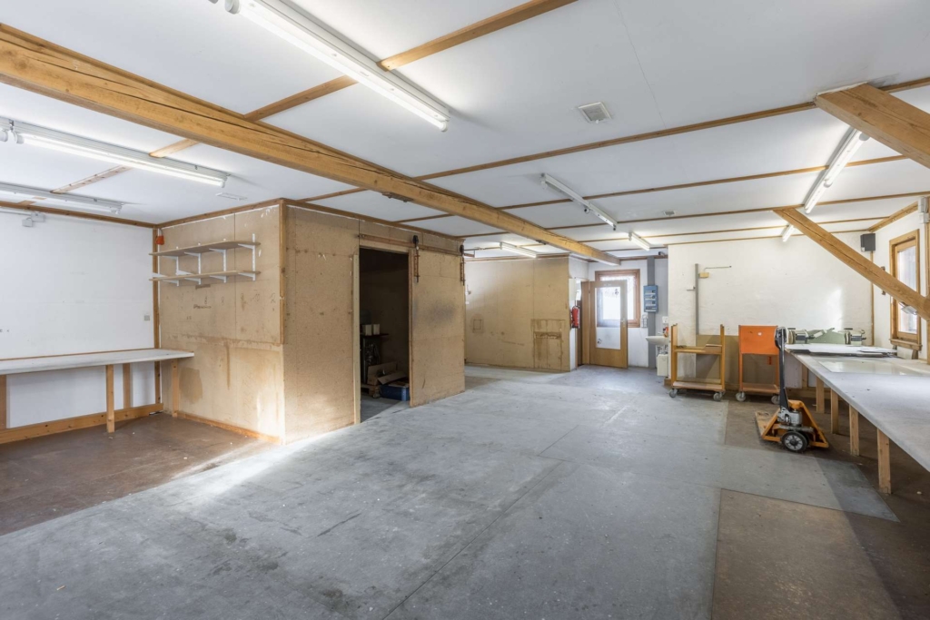 Eine geräumige, gut beleuchtete Werkstatt mit Holzbalken, weißen Wänden, Werkbänken, Regalen und einem zentralen geschlossenen Raum. Es gibt große Fenster, Leuchtstoffröhren und verschiedene Werkzeuge und Wagen im ganzen Raum.