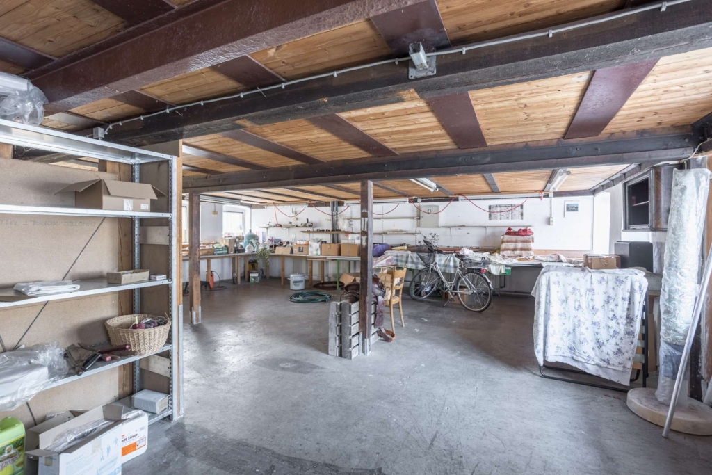 Eine geräumige, vollgestopfte Garage mit einer Holzdecke, Regalen mit verschiedenen Gegenständen auf der linken Seite, Fahrrädern, Arbeitstischen, Aufbewahrungsboxen und verschiedenen Werkzeugen, die überall verstreut sind. Durch kleine Fenster fällt natürliches Licht ein.