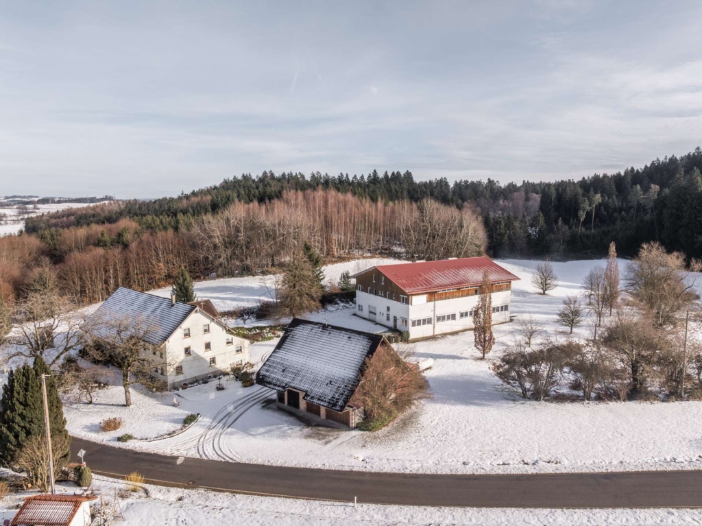Luftaufnahme von drei Gebäuden, darunter Häuser mit dunklen Dächern und ein größeres Gebäude mit einem roten Dach, umgeben von schneebedecktem Boden, Bäumen und einer Straße in einer ländlichen Winterlandschaft.