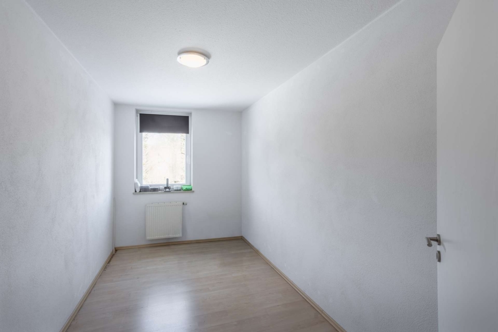 Ein kleiner, leerer Raum mit weißen Wänden, hellem Holzfußboden, einem einzigen Fenster mit einer dunklen Jalousie, einem Heizkörper unter dem Fenster und einer geschlossenen Tür, die rechts teilweise sichtbar ist.