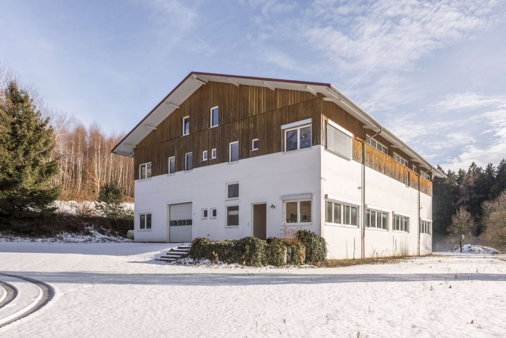 Ein großes zweistöckiges Gebäude mit weißem Untergeschoss und hölzernem Obergeschoss steht in einer verschneiten Landschaft, umgeben von Bäumen und einem teilweise bewölkten Himmel.