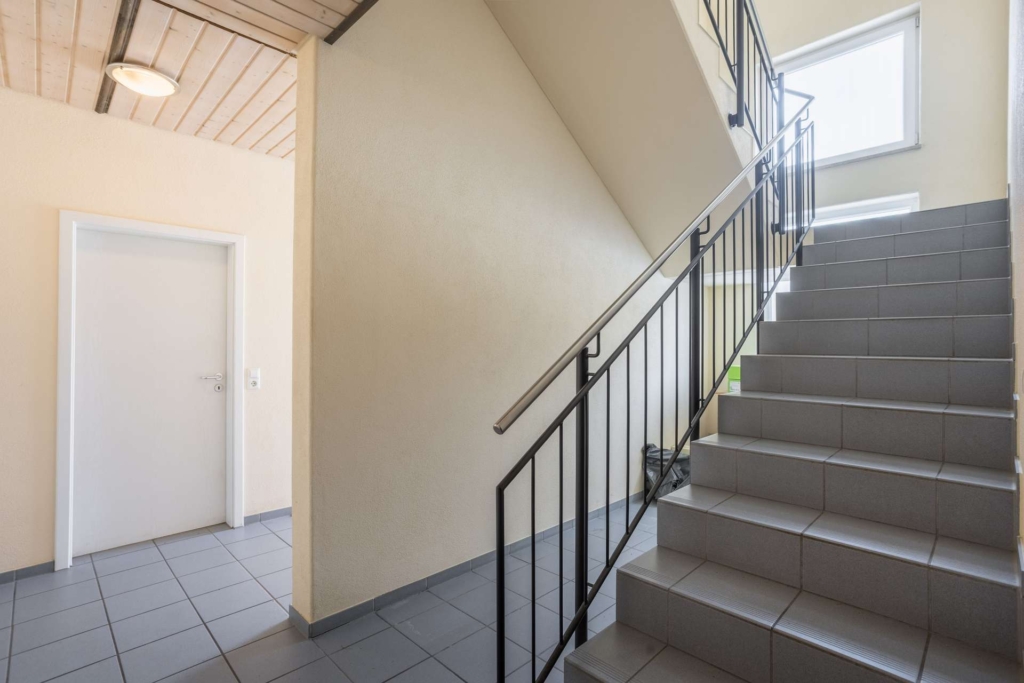 Eine moderne Innentreppe mit grau gefliesten Stufen und einem schwarzen Metallgeländer, daneben eine geschlossene weiße Tür und beigefarbene Wände, beleuchtet von natürlichem Licht aus einem Fenster über der Treppe.