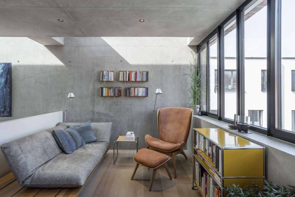 Modernes Wohnzimmer mit Betonwänden, großen Fenstern, einem grauen Sofa mit blauen Kissen, einem hellbraunen Ledersessel mit Ottomane, an der Wand befestigten Bücherregalen, einem gelben Schrank und einer Topfpflanze am Fenster.