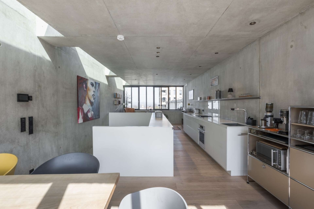 Moderne, minimalistische Küche mit Betonwänden und -decken, großen Fenstern, Einbaugeräten, offenen Regalen und modernen Möbeln. Ein farbenfrohes Porträt hängt an der Wand, und natürliches Licht erfüllt den Raum.