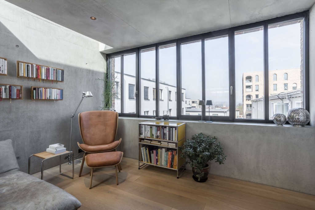 Modernes Wohnzimmer mit großen, bodentiefen Fenstern, hellem Holzfußboden, einem braunen Ledersessel, Bücherregalen voller Bücher, einer Topfpflanze und Blick auf die Nachbargebäude draußen.