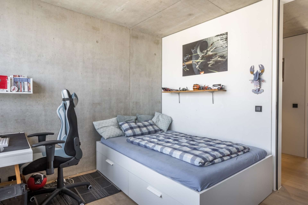 Ein modernes Schlafzimmer mit Betonwänden, einem Einzelbett mit blau kariertem Bettzeug, einem Spielesessel an einem Schreibtisch mit Büchern und einem Monitor sowie Regalen mit Modellautos und einem Poster mit Weltraummotiven über dem Bett.