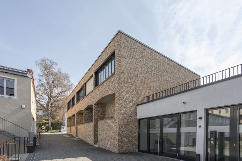 Ein modernes Gebäude mit hellbrauner Backsteinfassade und großen Fenstern mit bogenförmigen Öffnungen im Erdgeschoss, daneben ein weißer Bau mit Glastüren und Metallgeländern unter freiem Himmel.