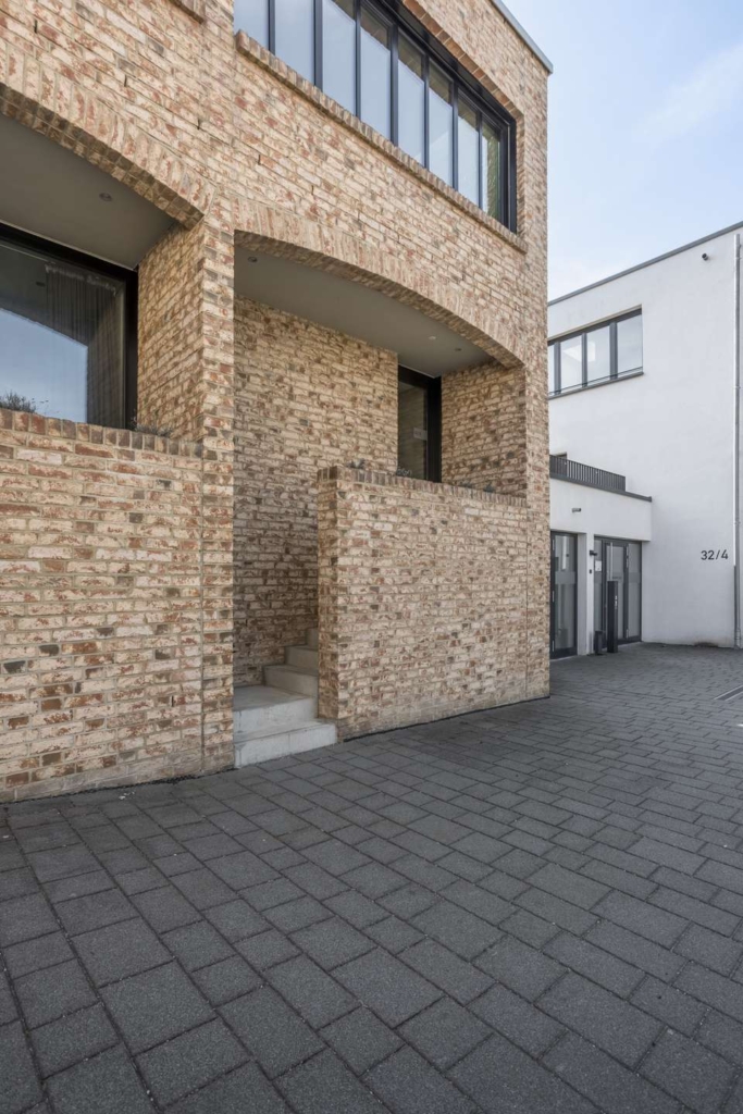 Modernes Gebäude mit hellbraunen Ziegelwänden, großen Fenstern und einer Betontreppe, die zu einem Eingang führt. Der Boden ist mit grauen Ziegeln gepflastert, und rechts ist ein weißes Gebäude zu sehen.