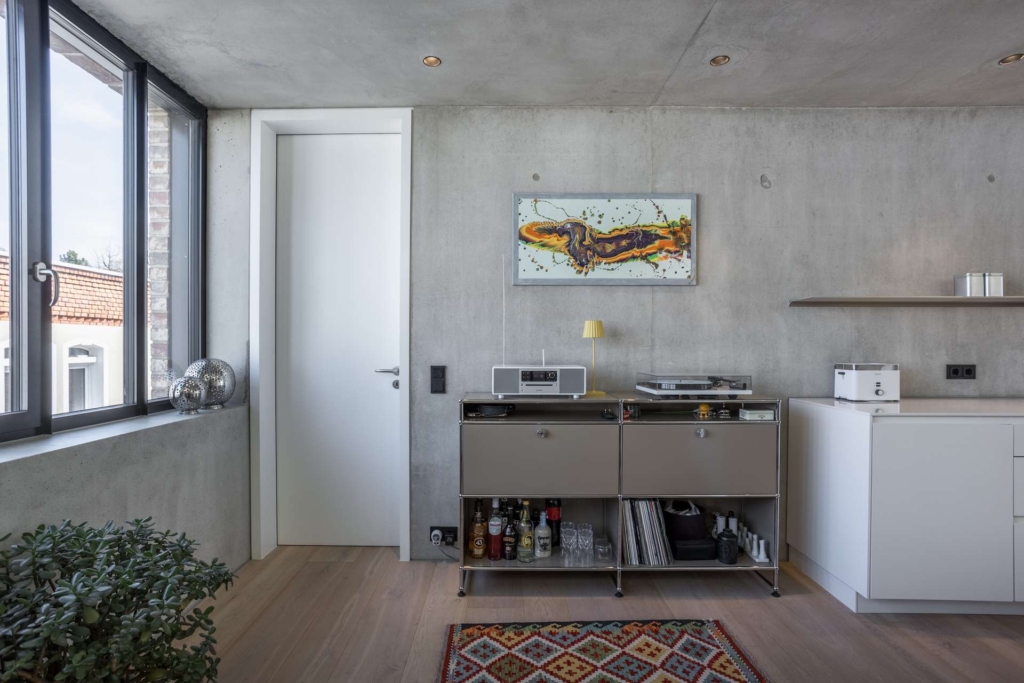 Ein moderner Raum mit Betonwänden, einer weißen Tür, zwei Schränken mit Getränken und Audiogeräten, einem bunten abstrakten Gemälde, großen Fenstern und einem gemusterten Teppich auf einem Holzboden.