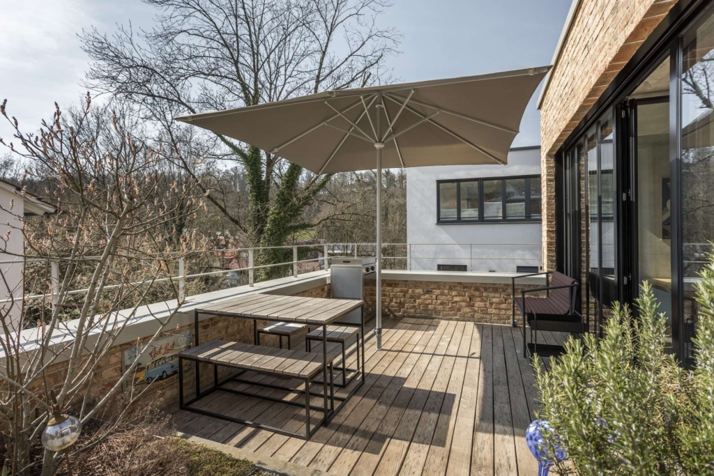 Eine moderne Außenterrasse mit Holzbelag bietet einen großen Sonnenschirm, einen Picknicktisch aus Metall mit Bänken, einen Loungesessel und einen Blick auf Bäume und nahe gelegene Gebäude. Glastüren öffnen sich vom Haus aus auf die Terrasse.