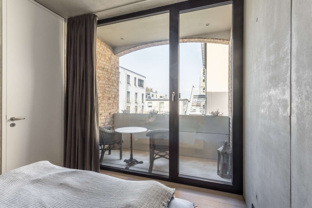 Ein modernes Schlafzimmer mit einer Glastür öffnet sich zu einem kleinen Balkon mit zwei Korbstühlen, einem runden weißen Tisch und einer Laterne. Der Balkon ist von Backsteinmauern umrahmt und bietet einen Blick auf die benachbarten Gebäude.