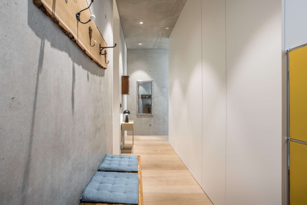 Minimalistischer Flur mit hellem Holzboden, weißen Wänden, einem großen Wandspiegel, einem kleinen Waschbecken, gelben Schränken, einer Holzbank mit blauen Kissen und Kleiderhaken an einer Betonwand. Helles und modernes Interieur.