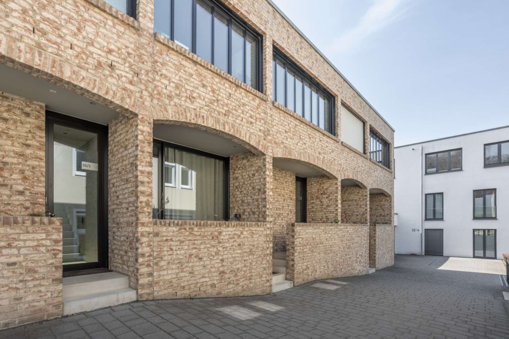 Ein modernes Wohnhaus aus Backstein mit gewölbten Eingängen und großen Fenstern, mit einem gepflasterten Gehweg und einem weißen Gebäude im Hintergrund, das bei klarem Himmel zu sehen ist.