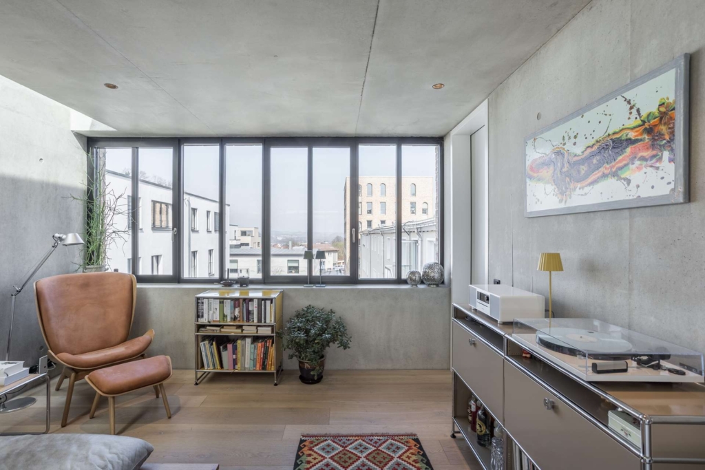 Ein modernes, minimalistisches Wohnzimmer mit großen Fenstern, einem braunen Sessel, einem Bücherregal, einem Plattenspieler auf einer Metallkonsole, einer kleinen Pflanze und einem abstrakten Kunstwerk an einer Betonwand. Draußen sind städtische Gebäude zu sehen.