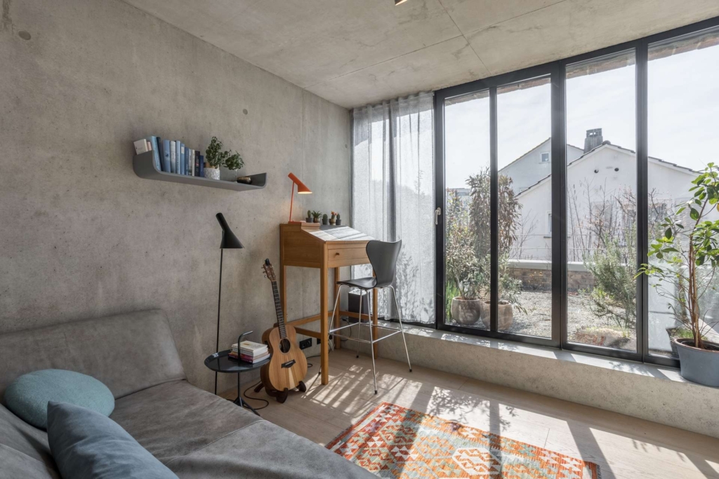 Ein heller, moderner Raum mit Betonwänden, einem Sofa, einem gemusterten Teppich, einem Holzschreibtisch neben großen Glastüren, einer Gitarre, Pflanzen und Büchern in einem schwebenden Regal. Das Sonnenlicht strömt von der Terrasse nach draußen.