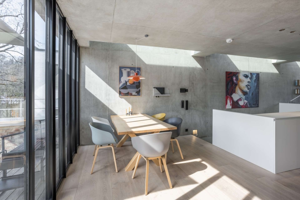 Ein moderner Essbereich mit einem Holztisch, vier bunten Stühlen, großen, raumhohen Fenstern, Betonwänden und zeitgenössischen Kunstwerken. Sonnenlicht strömt herein und erzeugt helle Muster an den Wänden und auf dem Boden.