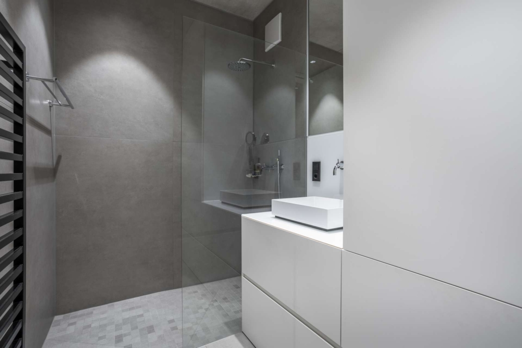 Modernes, minimalistisches Badezimmer mit grau gefliesten Wänden, einer gläsernen Dusche und einem weißen Waschtisch mit einem quadratischen Waschbecken. Der Raum ist sauber, aufgeräumt und gut beleuchtet und verfügt über einfache, moderne Armaturen.